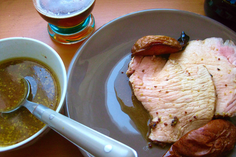 Slices of pork and pears on a plate beside a small bowl of mustard sauce with a spoon and a glass of beer.